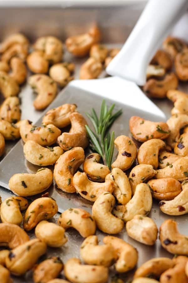 Rosemary cashews on a metal baking pan with a spatula and sprig of fresh rosemary.