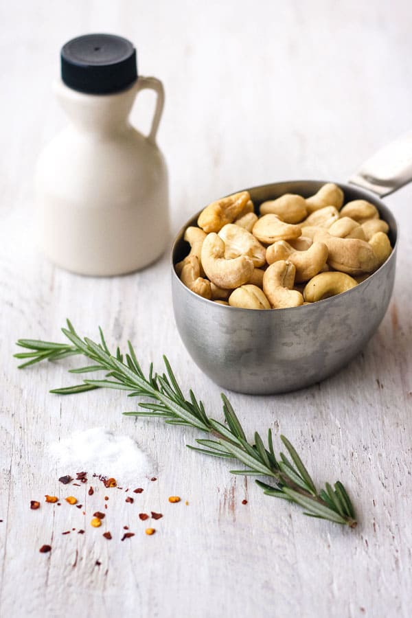 Raw cashews in a metal measuring cup, a small bottle of maple syrup, fresh sprig of rosemary and seasonings on a white wood table.