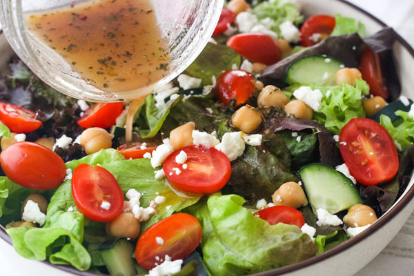 Italian dressing pouring from a mason jar onto a green salad with tomatoes and feta.