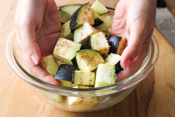 Hands tossing seasoned eggplant chunks.