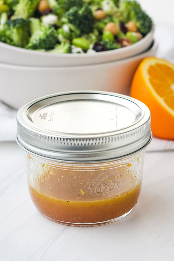 Salad dressing in a small mason jar in front of a bowl of salad and half a fresh orange.