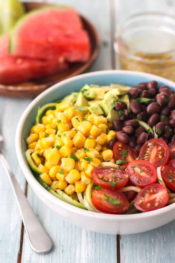 A bowl of zucchini noodle salad on a table with watermelon slices and vinaigrette