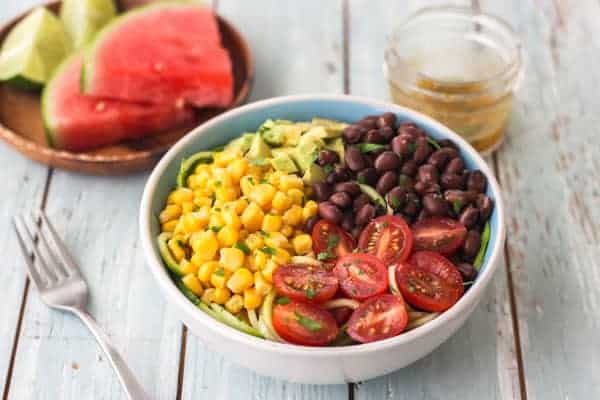 A blue bowl of zucchini noodle salad on a table with a plate of sliced watermelon.