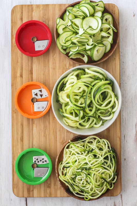3 types of zucchini noodles in bowls next to their corresponding blade attachment.