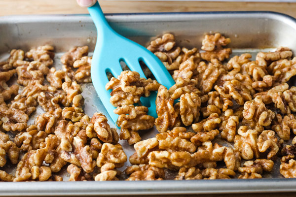 A blue spatula stirring coated walnuts on a sheet pan.