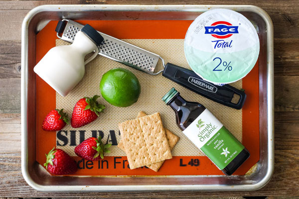 Yogurt container, strawberries, a lime, graham cracker and vanilla on a baking sheet.