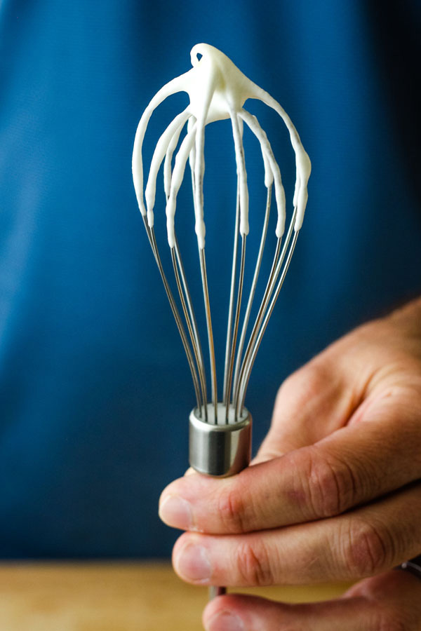 A man's hand holding a whisk covered in whipped cream with the tip folding over.