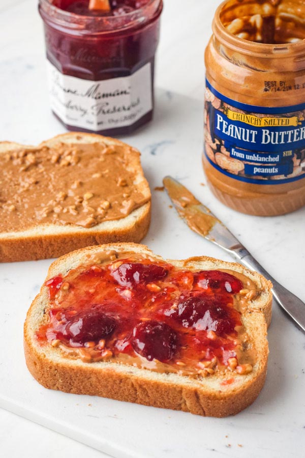 A cutting board with bread slices spread with crunchy peanut butter and strawberry preserves.