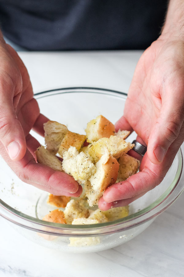 Hands tossing bread cubes and seasoning in a bowl.