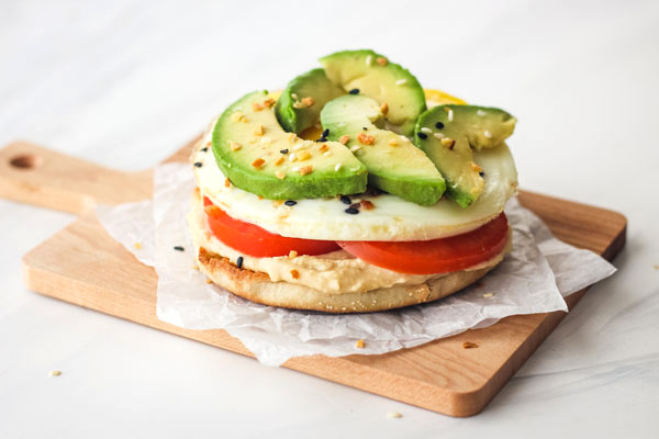 Small cutting board with an english muffin topped with hummus, egg, and avocado slices.