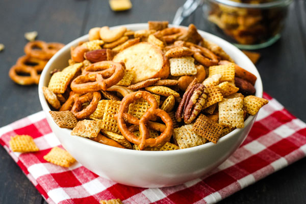 Snack mix in a white bowl on a red checkered napkin.