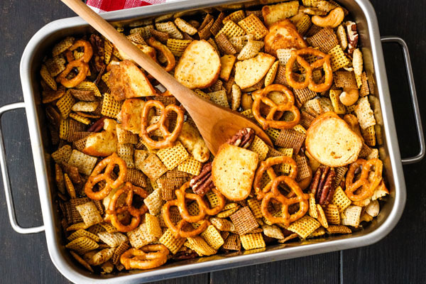 Snack mix in a small roasting pan with a wooden spoon.