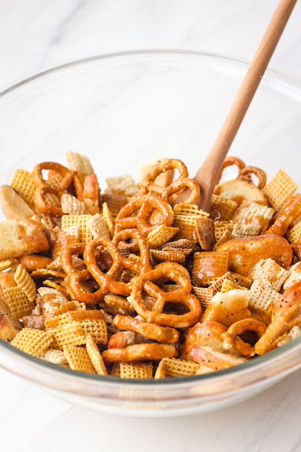 Chex mix ingredients in a glass bowl with a wooden spoon.