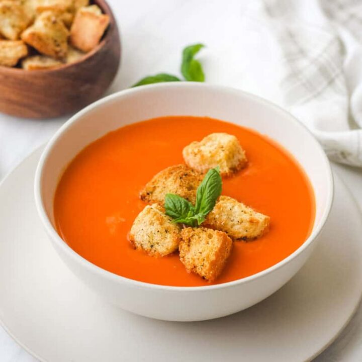 Bowl of red pepper soup topped with croutons and basil leaves.