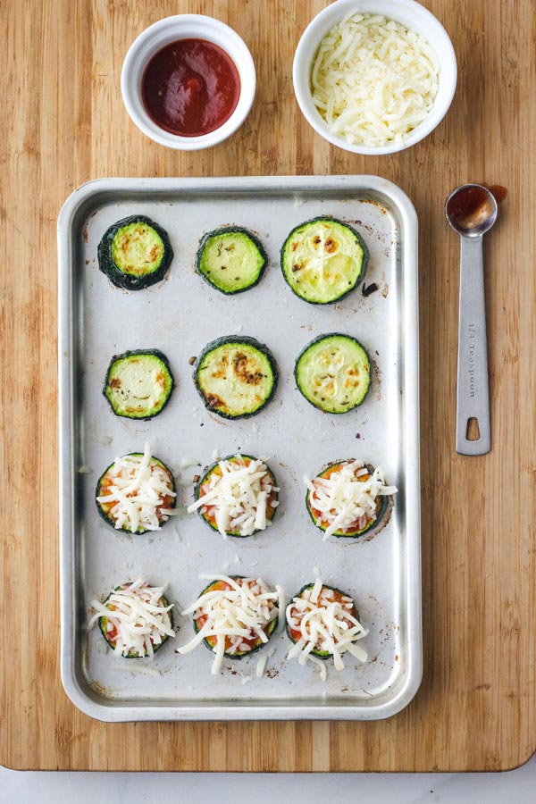 Broiled zucchini on a cutting board with bowls of pizza sauce and shredded mozzarella cheese.