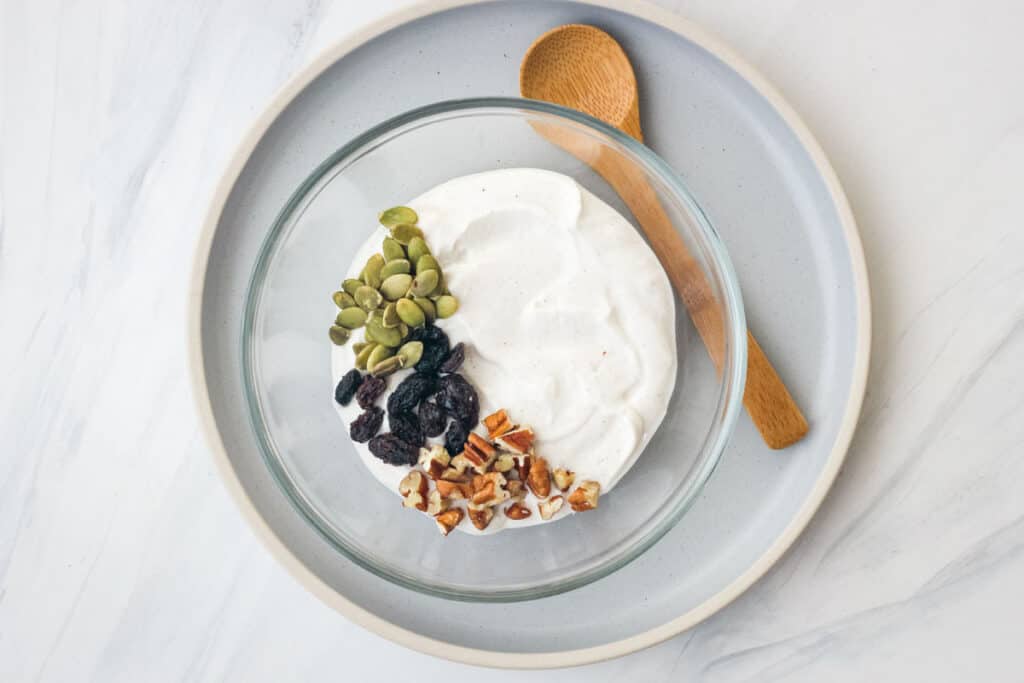 Glass bowl of yogurt topped with pepitas, nuts, and raisins.