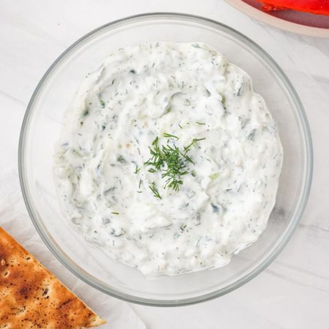 A small glass bowl of tzatziki sauce with veggies and pita bread.