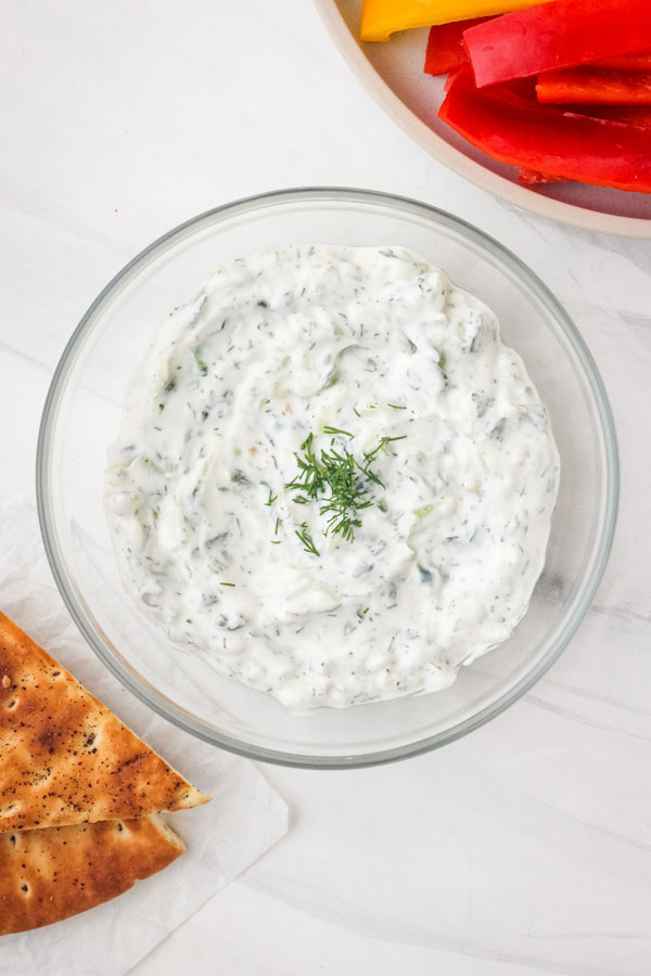 A small glass bowl of tzatziki sauce with veggies and pita bread.