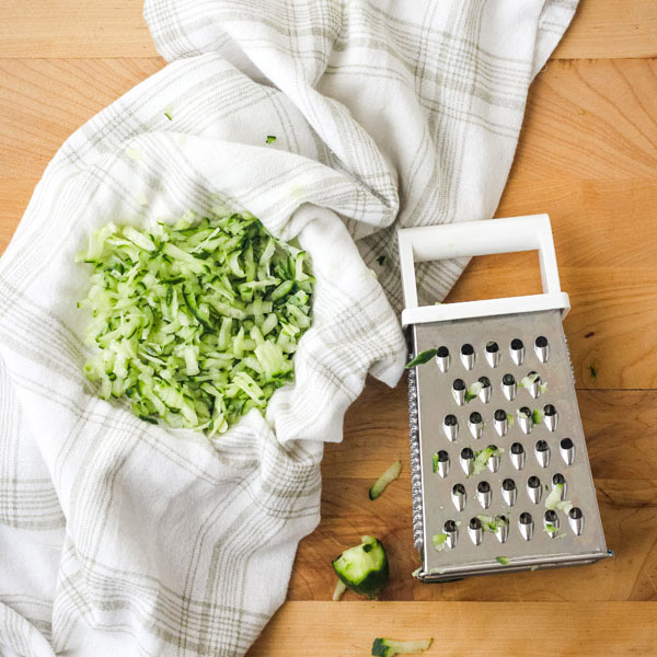 Shredded cucumber in white kitchen towel next to a grater.