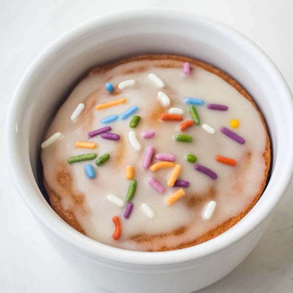 A white ramekin filled with cake and topped with glaze and rainbow colored sprinkles.