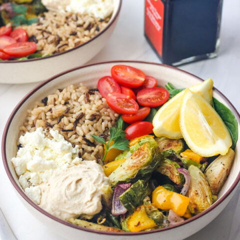 Two tan bowls with grains and roasted vegetables with a bottle of balsamic vinegar.