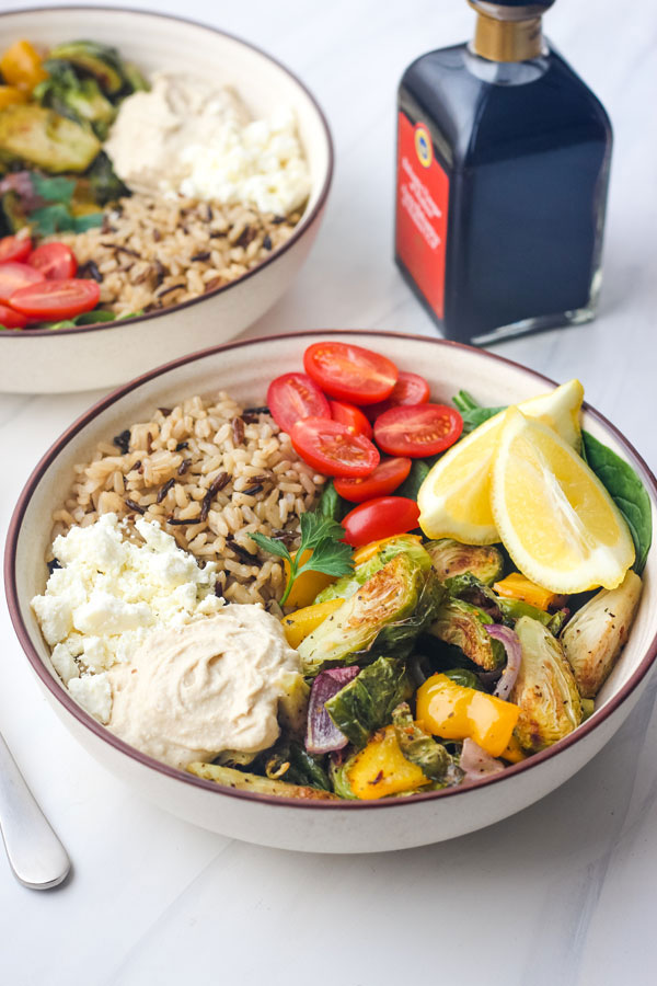 Two tan bowls with grains and roasted vegetables with a bottle of balsamic vinegar. 