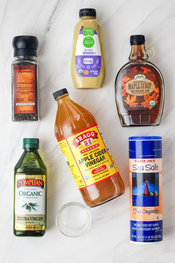 Containers of olive oil, vinegar, maple syrup, mustard, salt, and pepper on a table.