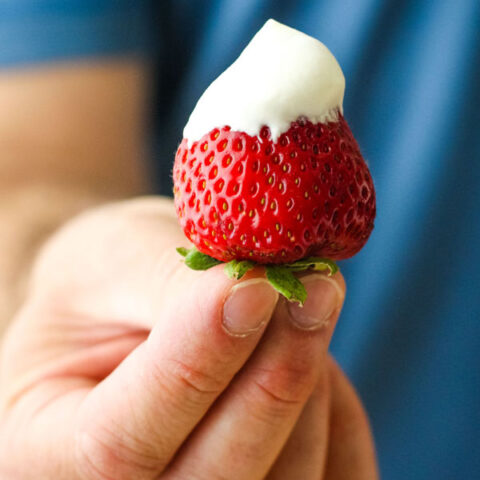 Man in a blue shirt holding a strawberry dipped in whipped cream.