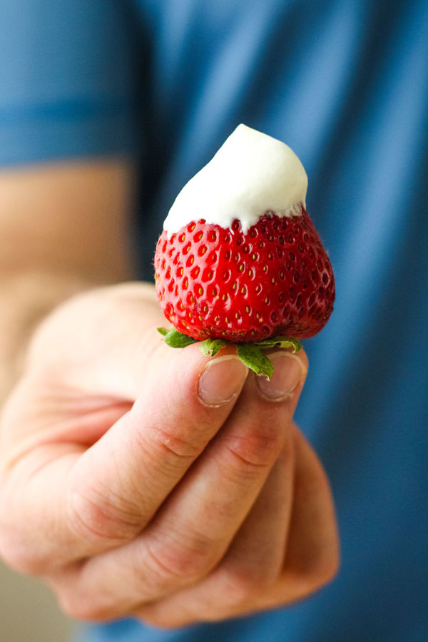 Man in a blue shirt holding a strawberry dipped in whipped cream.