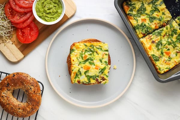 Plate with half a bagel surrounded by a cutting board with sliced tomato and guacamole, pan of baked eggs, and toasted bagels.
