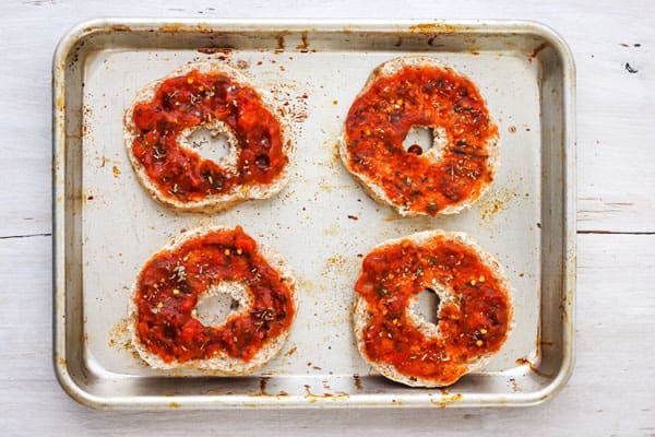 Sauce-topped bagel halves on sheet pan.