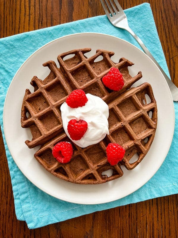 Plate with a chocolate waffle topped with a dollop of whipped cream and fresh raspberries.