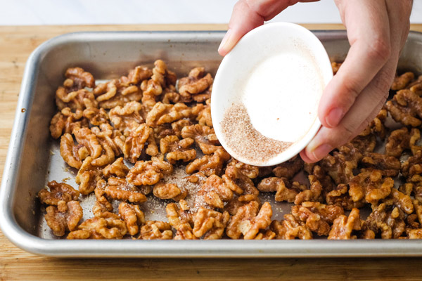 A hand shaking cinnamon sugar over toasted walnuts on a sheet pan.