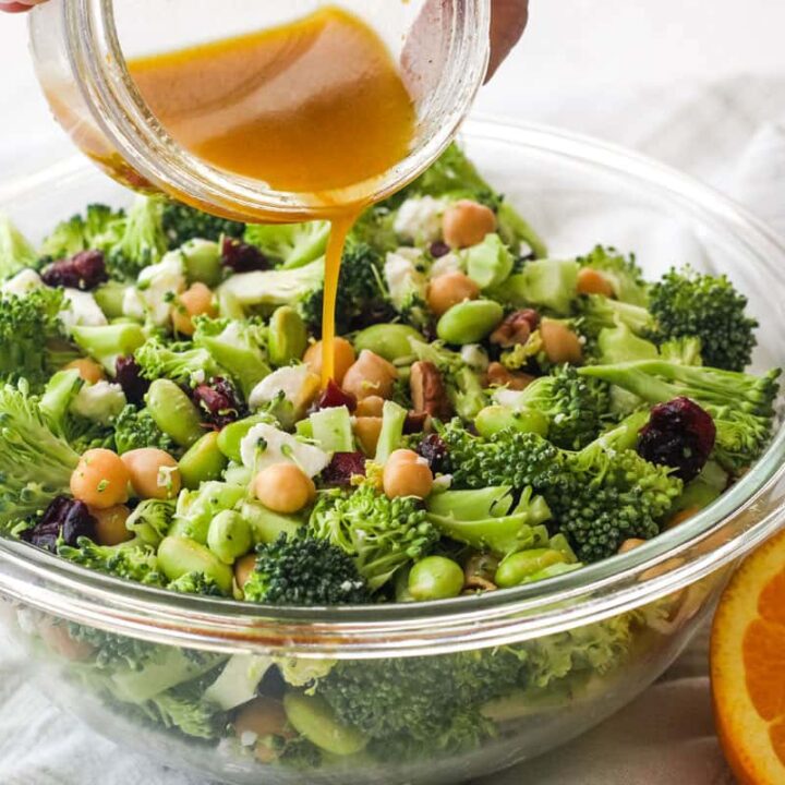 Hand pouring orange vinaigrette over a bowl of broccoli salad.