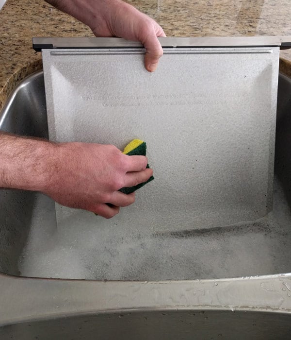 Man washing a crumb tray in a sink of soapy water with a sponge.