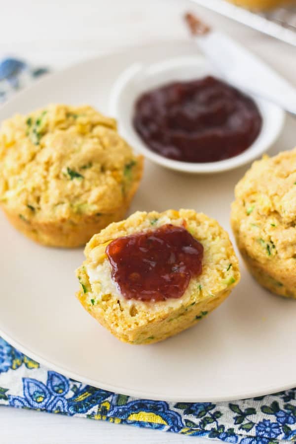 Cornbread zucchini muffins sliced in half on a plate and spread with butter and strawberry preserves.