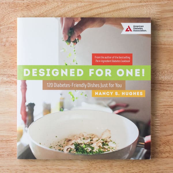 Designed For One cookbook cover with hand sprinkling herbs into a skillet.