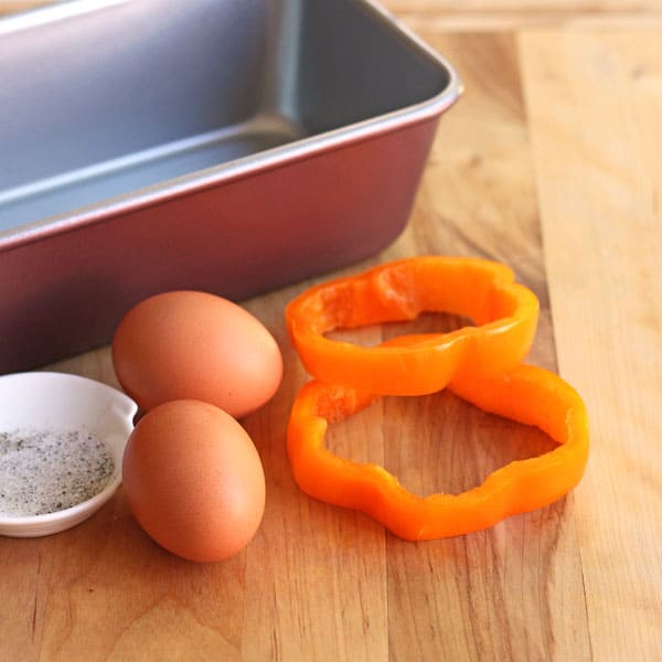 A loaf pan, eggs, and two bell pepper rings on a cutting board.