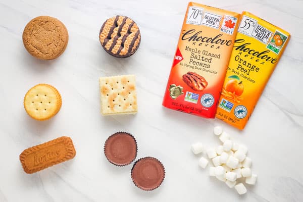 Cookies, crackers, peanut butter cups, mini marshmallows, and chocolate bars arranged on a counter.