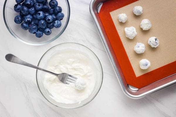 Fork scooping into a bowl of yogurt next to a bowl of fresh blueberries and a lined pan with yogurt-coated blueberries.