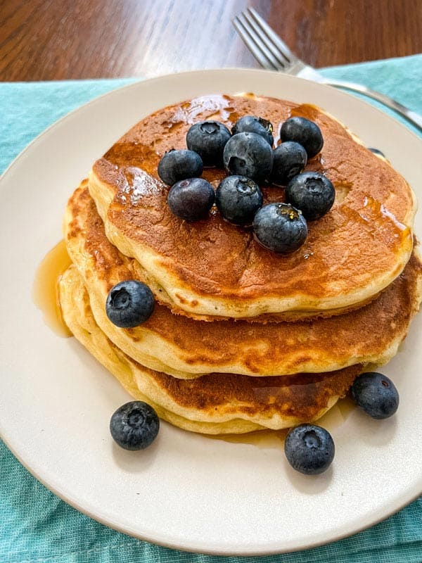 Stack of three fluffy pancakes with a pile of fresh blueberries and maple syrup.