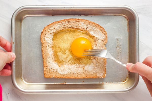 Fork poking egg yolk in slice of bread.