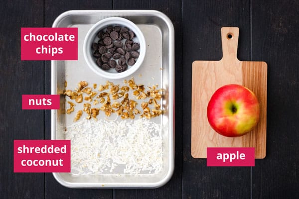 Apple on a small cutting board and small sheet pan with coconut, nuts, and ramekin of chocolate chips.