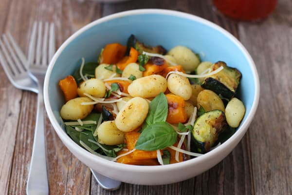 A blue bowl with roasted gnocchi and vegetables on a wooden table.