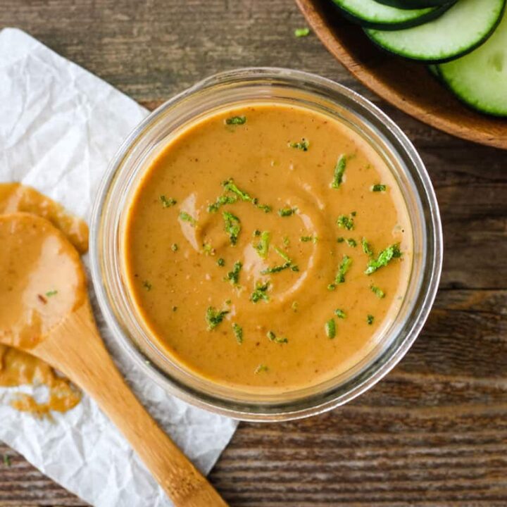 Small jar of peanut sauce with lime zest next to wooden bowl of raw veggies.