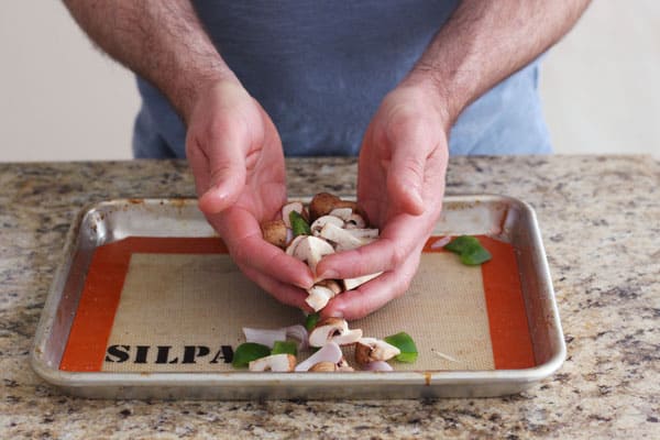 Hands tossing vegetables with oil on a baking sheet.