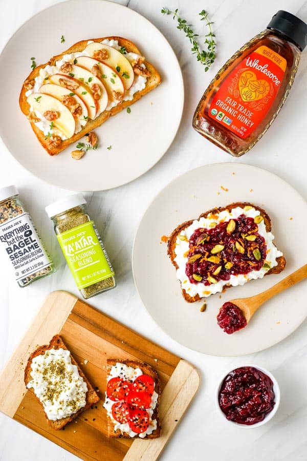 Plates of cottage cheese toasts, jars of seasoning, honey, and fresh herbs on a table.